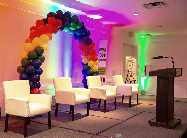 Rainbow balloon arch and white chairs on a lit stage beside a podium