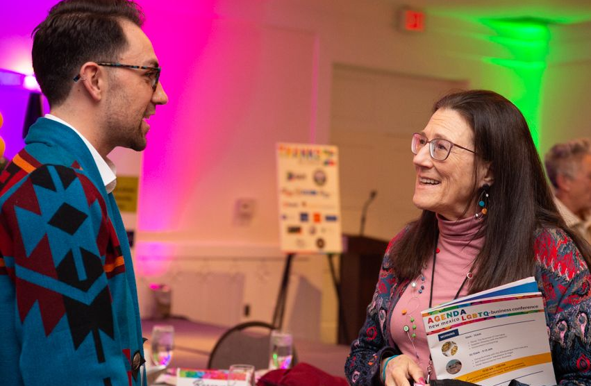 Two people talking at a conference table under pink and green lights, one holding a brochure.
