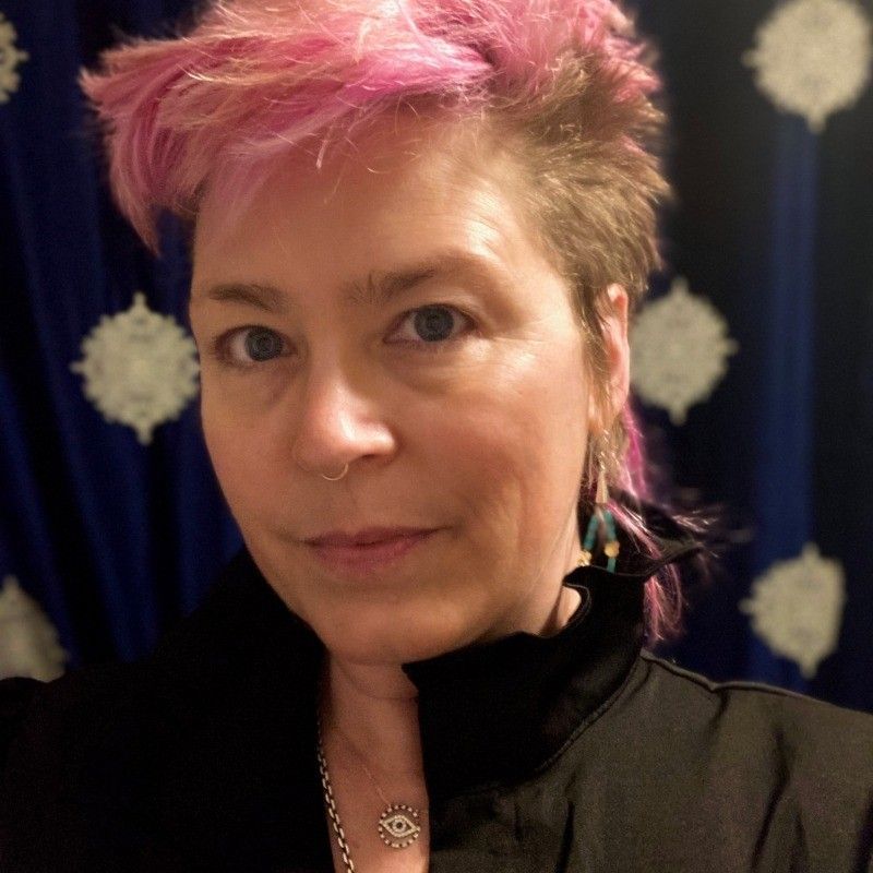 Portrait of a person with short pink hair, wearing a black top against a dark blue patterned background
