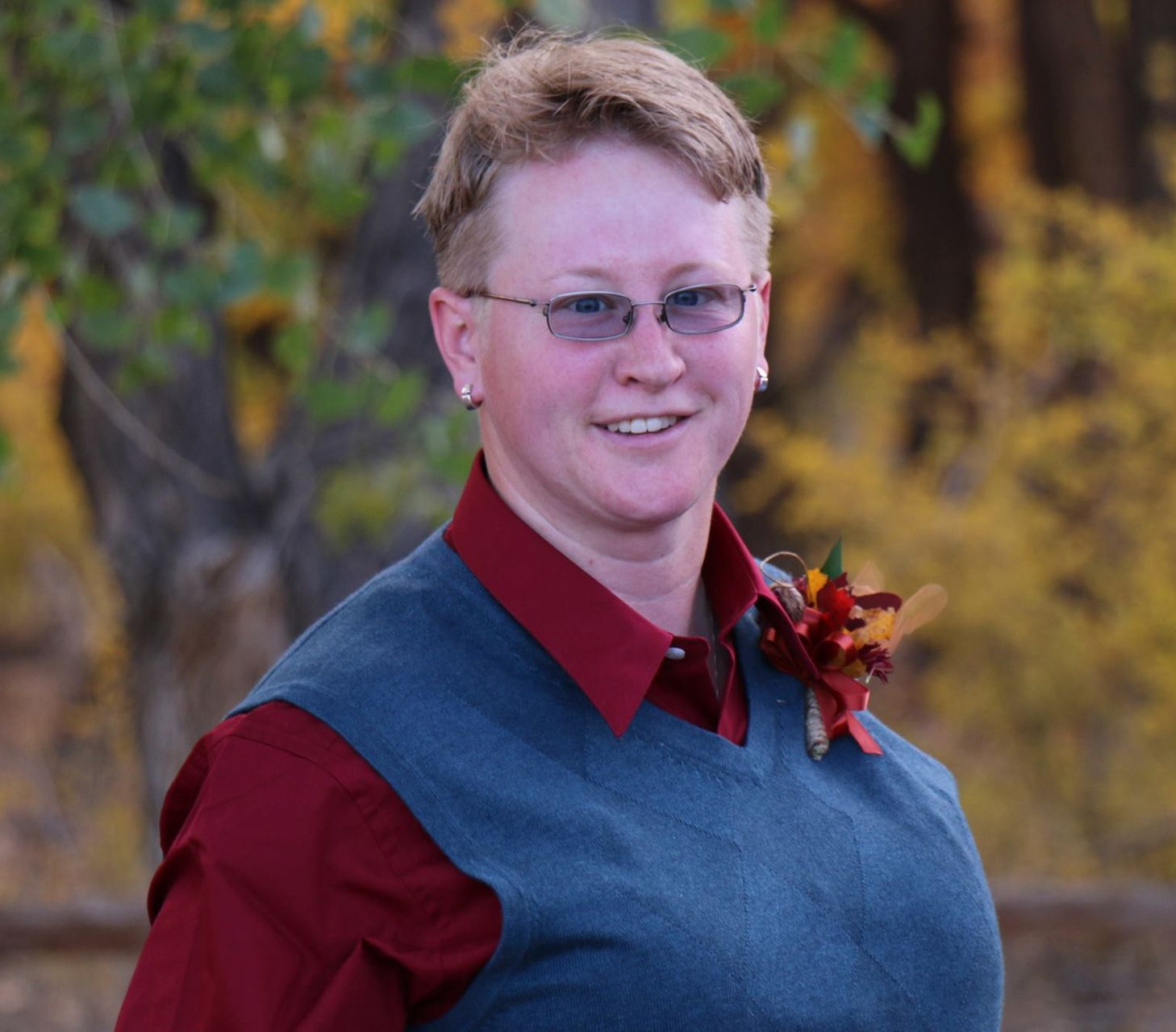 Smiling person in glasses and a blue vest with red shirt, standing outdoors against a blurred autumn background
