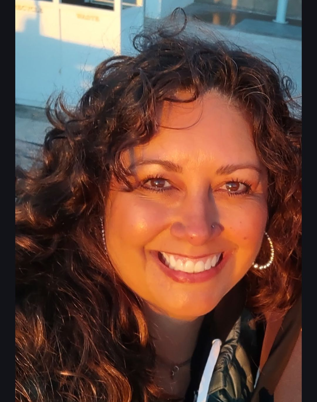 Smiling woman with curly hair in warm sunlight, wearing hoop earrings and a jacket
