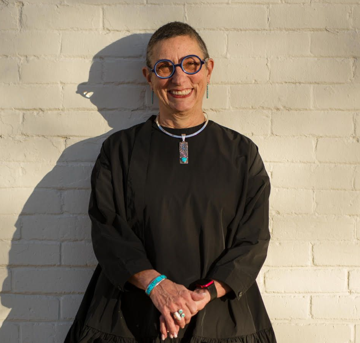 Smiling person in black clothing and glasses standing against a white brick wall in sunlight