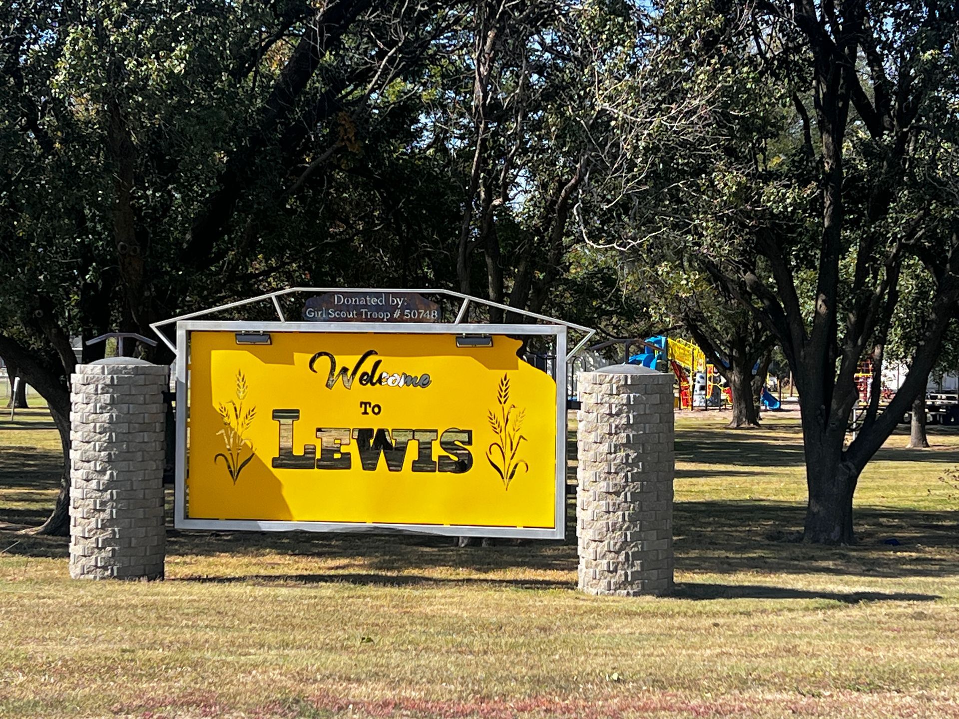 A yellow sign that says welcome to lewis is in a park surrounded by trees.