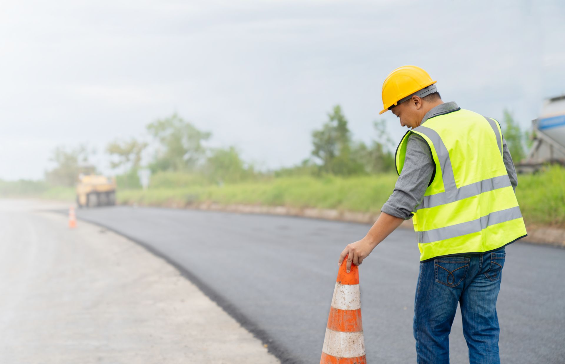 A construction worker is standing next to a traffic cone on the side of a road.