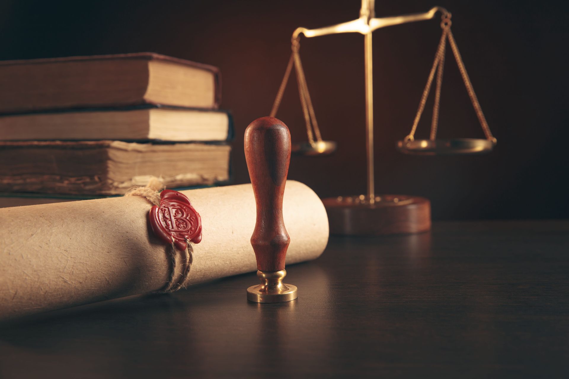 A stack of books, a rolled parchment with a red wax seal, a stamp, and a gold scale of justice on a dark wooden table.