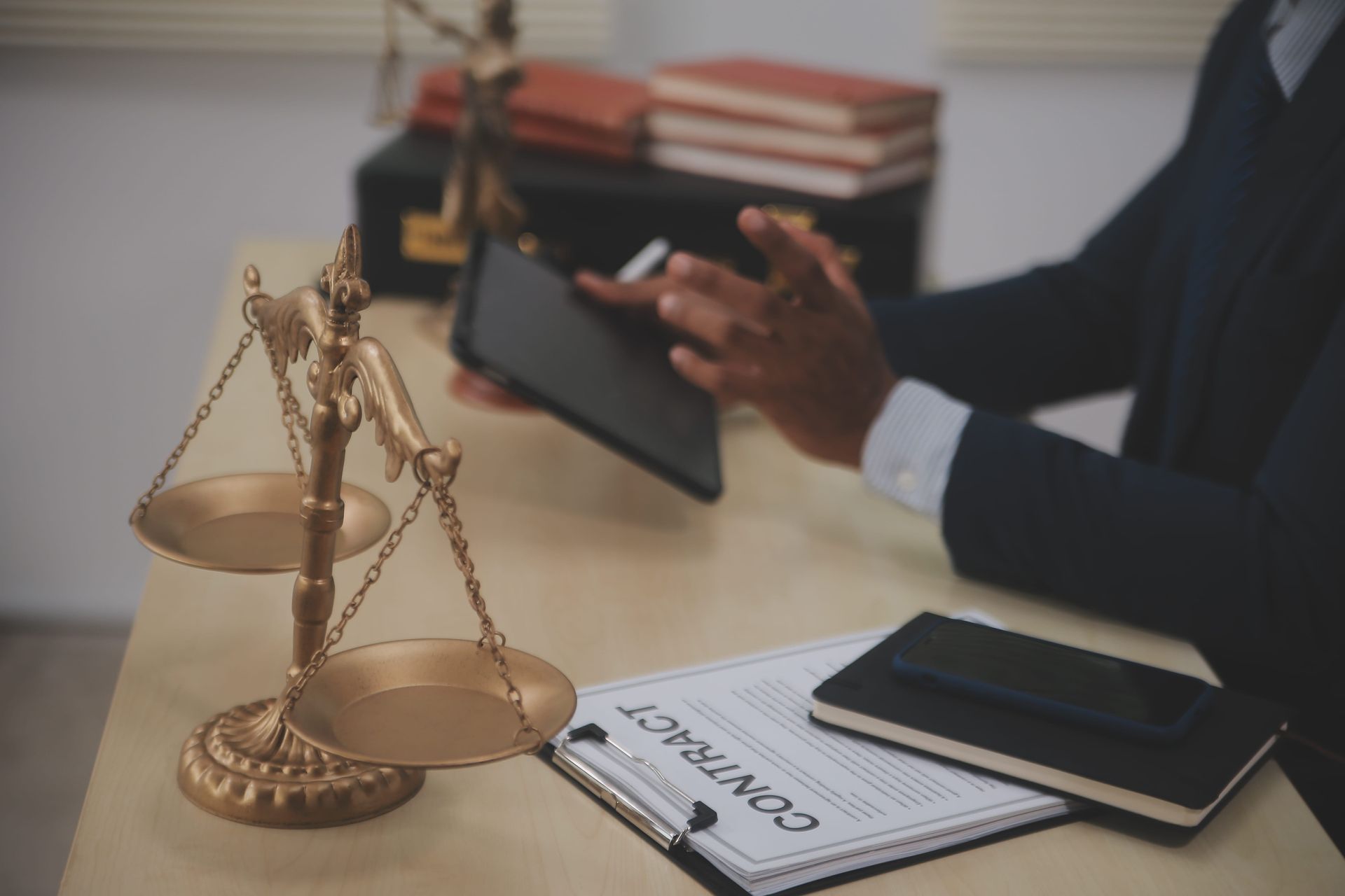 A person in a suit reviews a tablet at a desk with a scale of justice, a contract, and a black notebook.