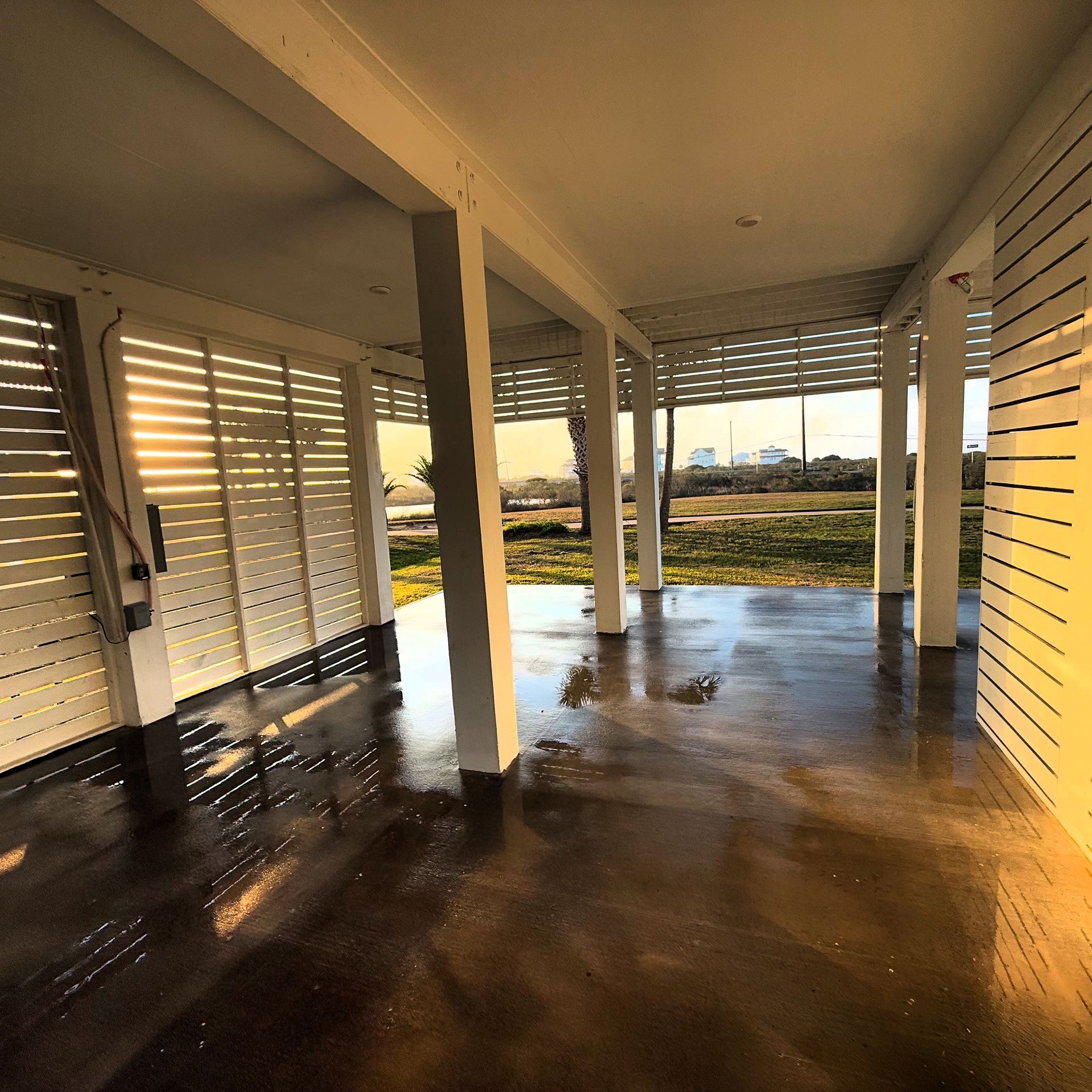 Covered porch with wet floor, white columns, and slatted walls overlooking a grassy waterfront at sunset