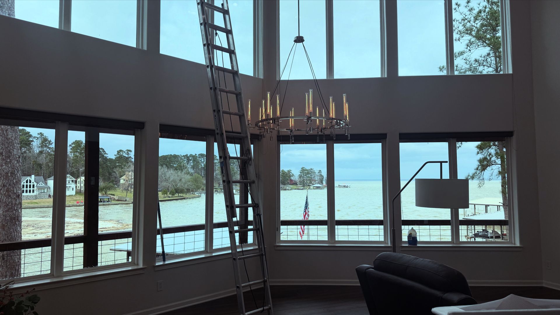 Modern living room with floor-to-ceiling windows overlooking water, a chandelier, and a ladder near the ceiling.
