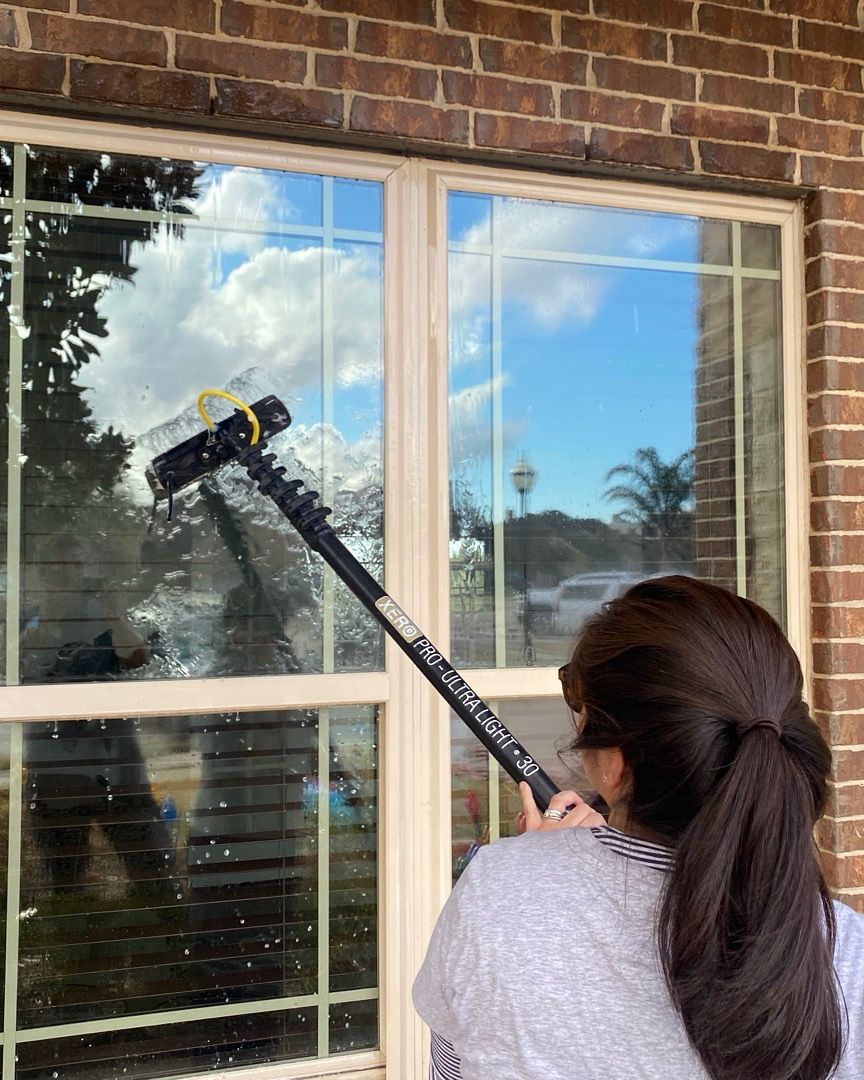 Person using a long-handled window cleaner on a brick house window with reflected sky and clouds