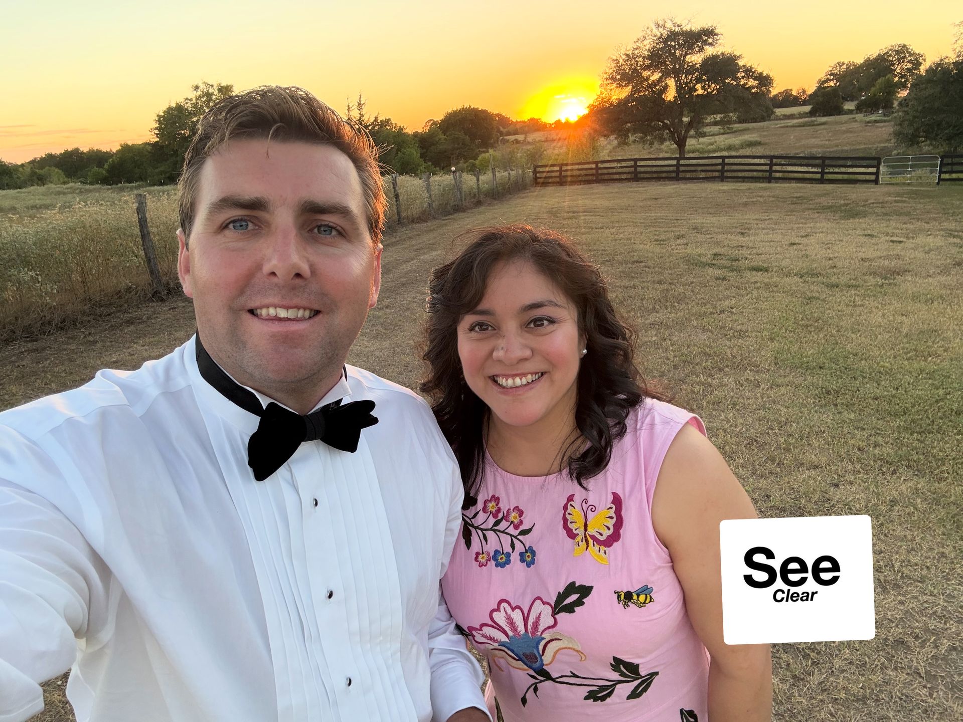Two people smiling outdoors at sunset, wearing formal and floral attire.