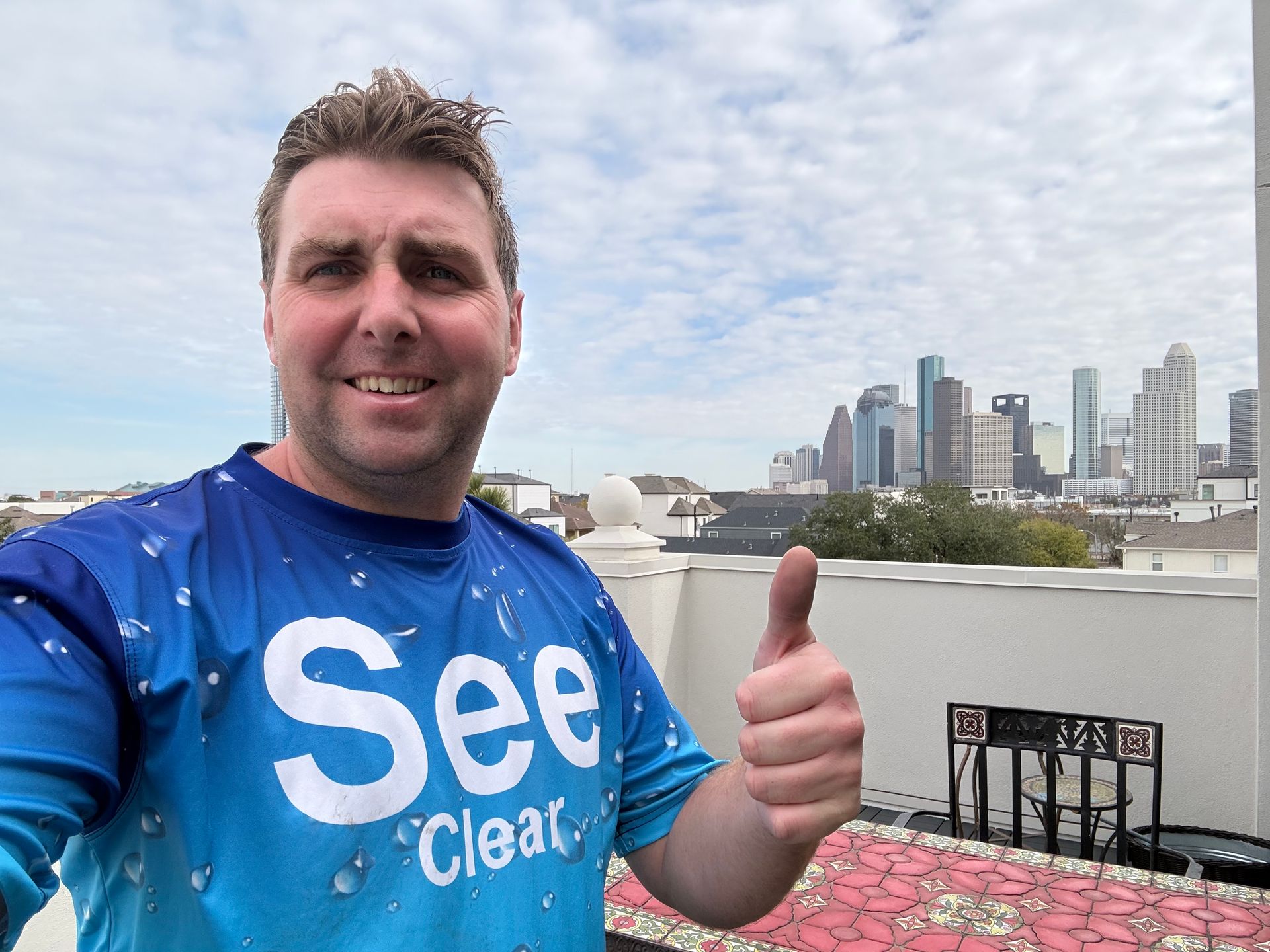 Man in blue “See” shirt giving thumbs up with city skyline and harbor in background