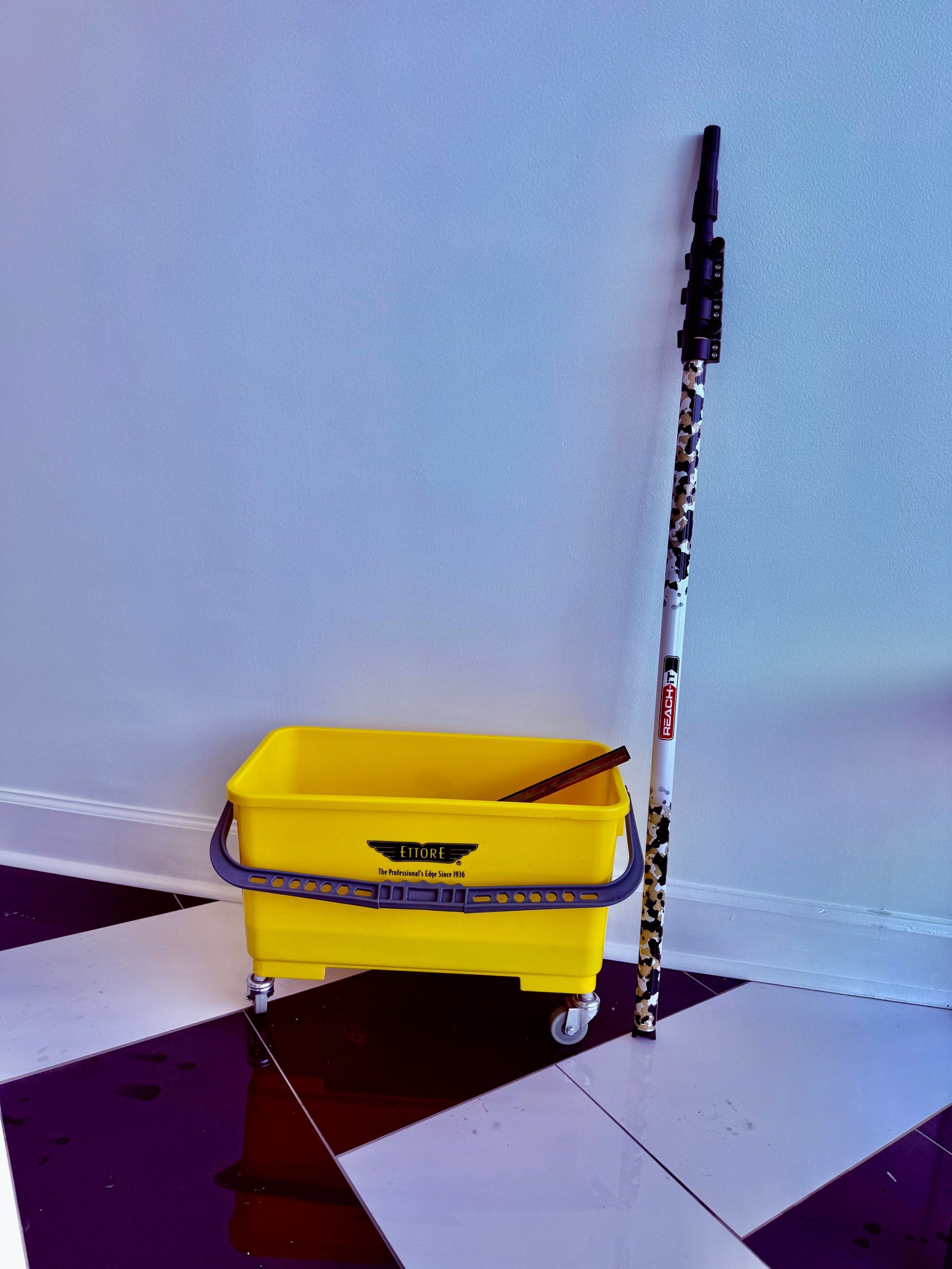 Yellow mop bucket and mop leaning against a wall on a tiled floor.