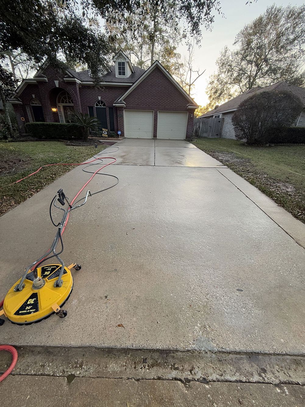 Driveway with a yellow pressure washer hose reel in the foreground, leading to a house and garage at sunset