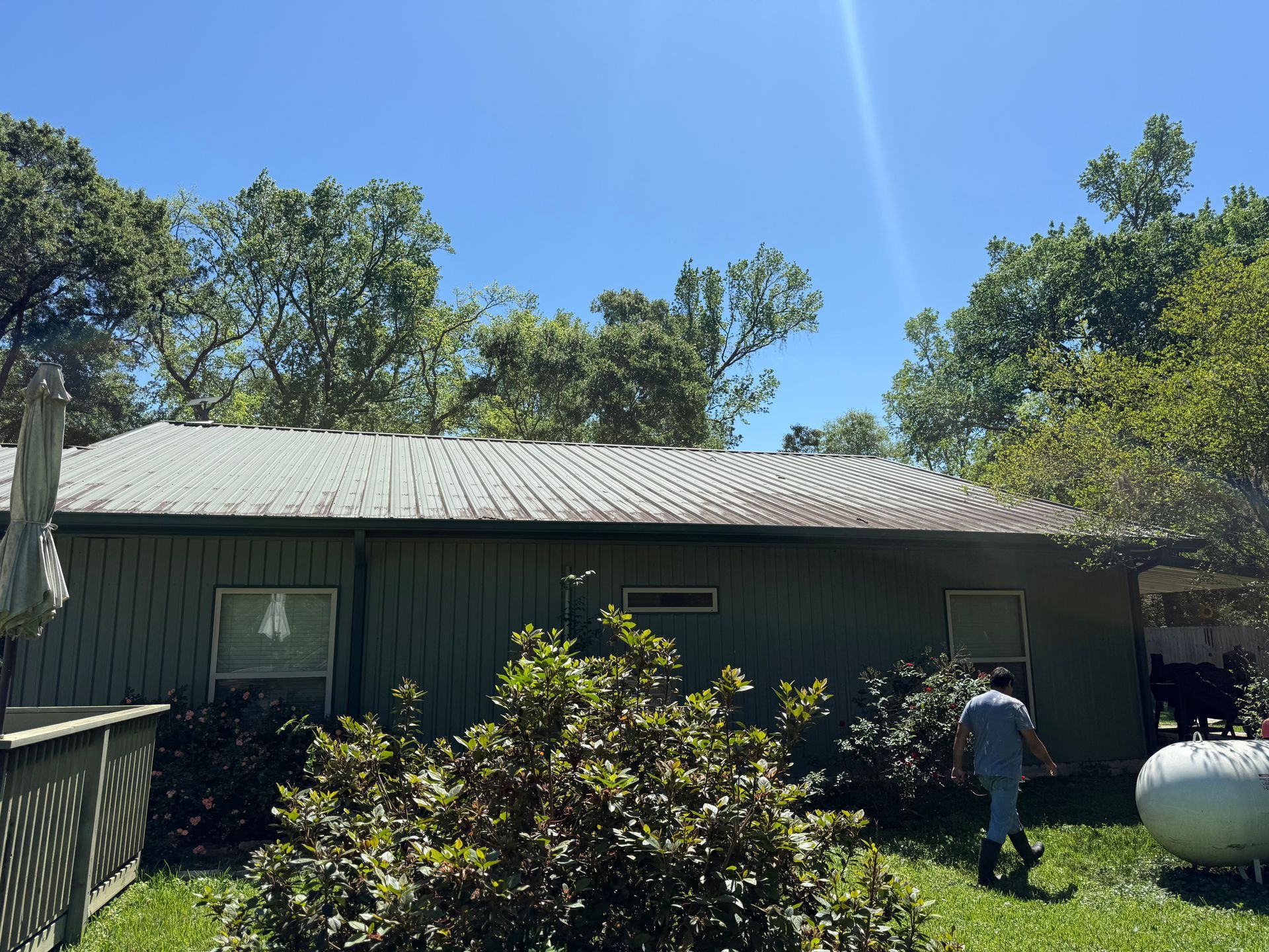 Single-story gray house with trees under a bright blue sky; a person stands near a large white tank.