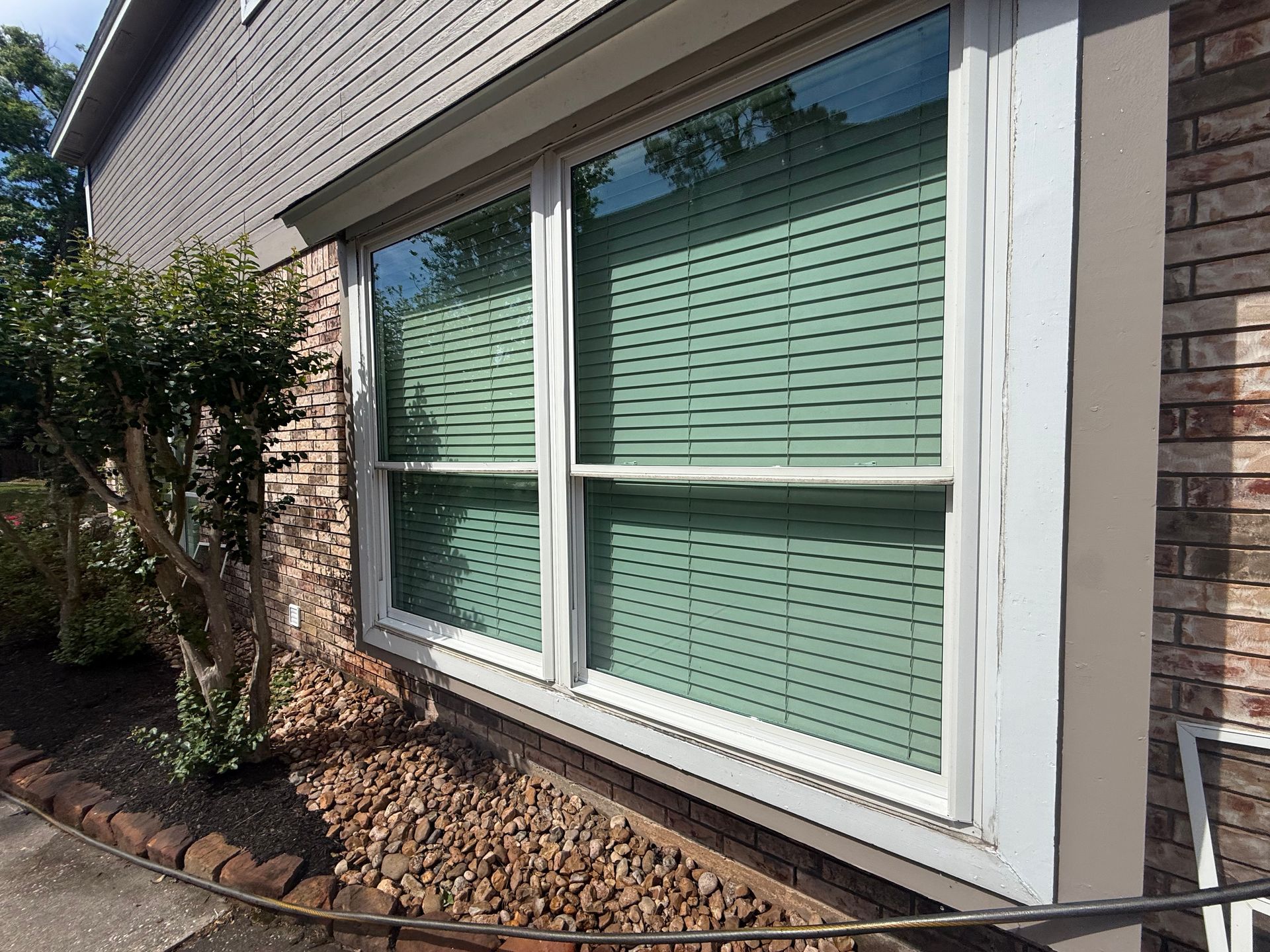 White-trimmed bay window with green blinds on a brick house, bordered by landscaping rocks and shrubs