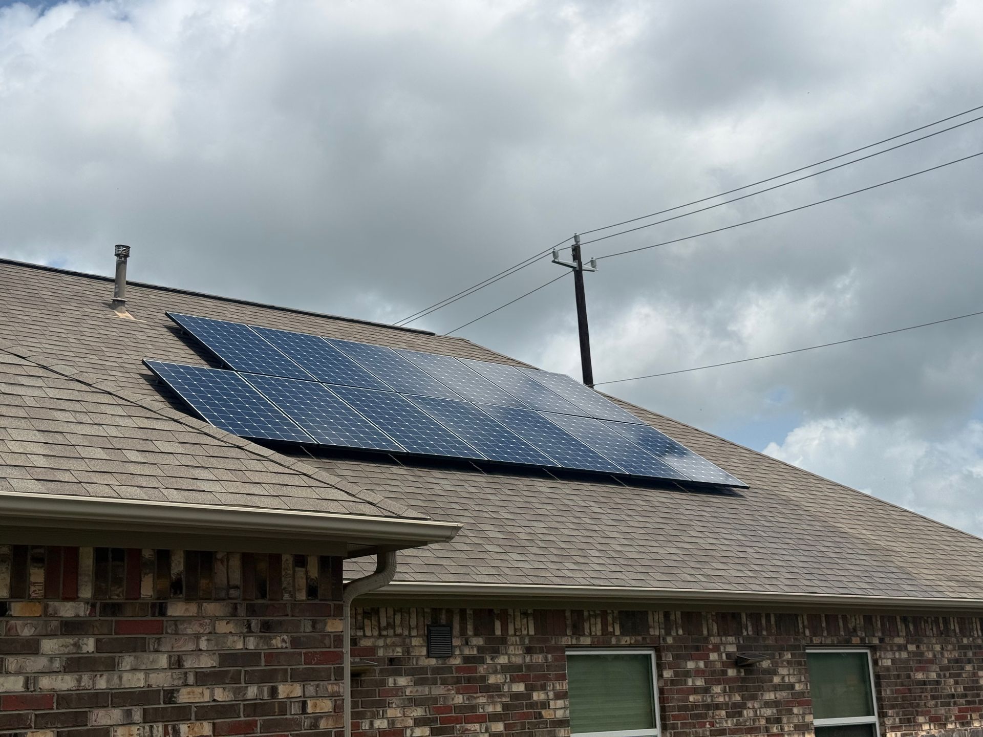 Solar panels mounted on a shingled roof under a cloudy sky, with brick walls and power lines visible.