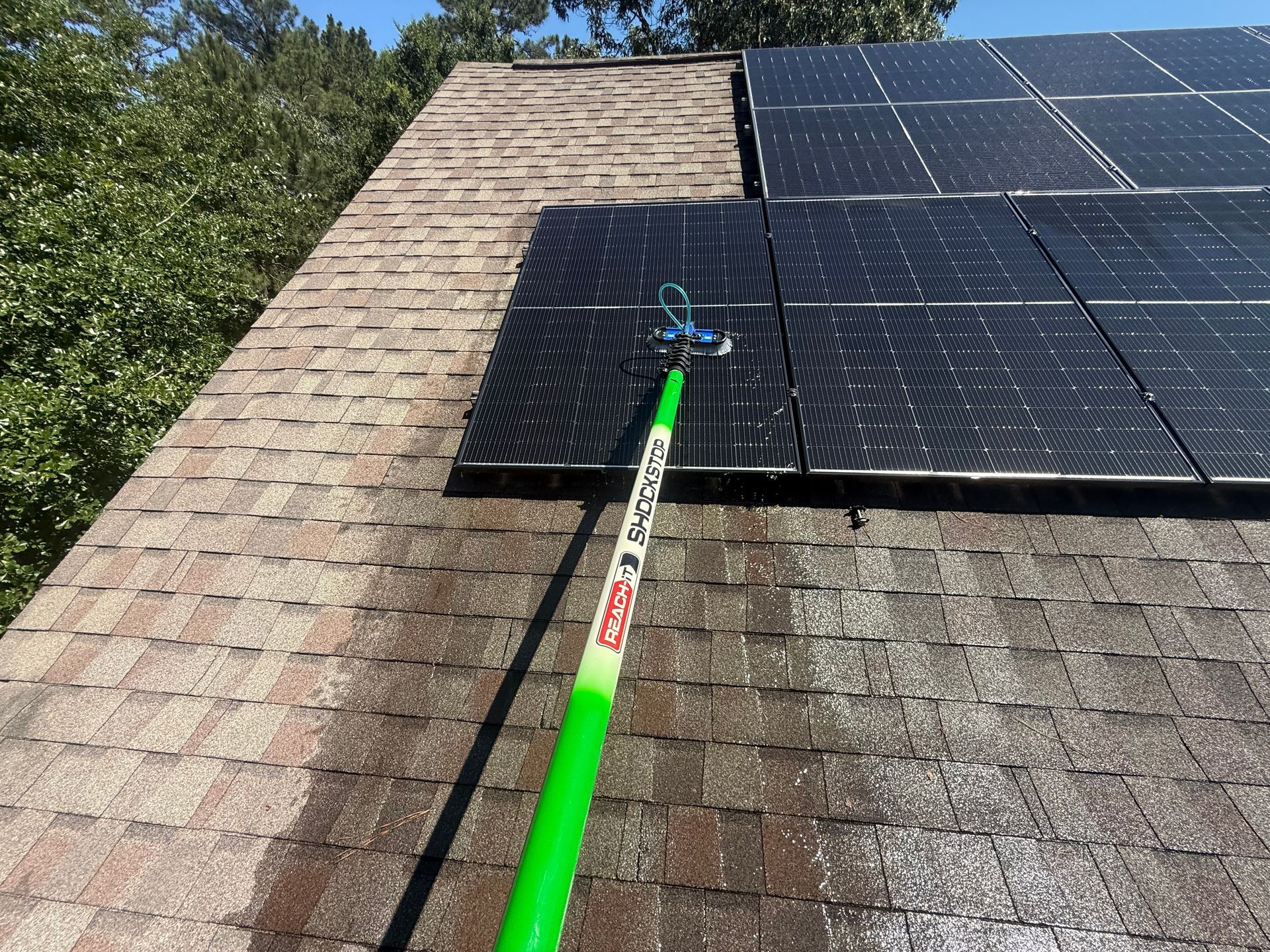 Solar panels on a shingled roof being cleaned with a green-handled brush on a pole.
