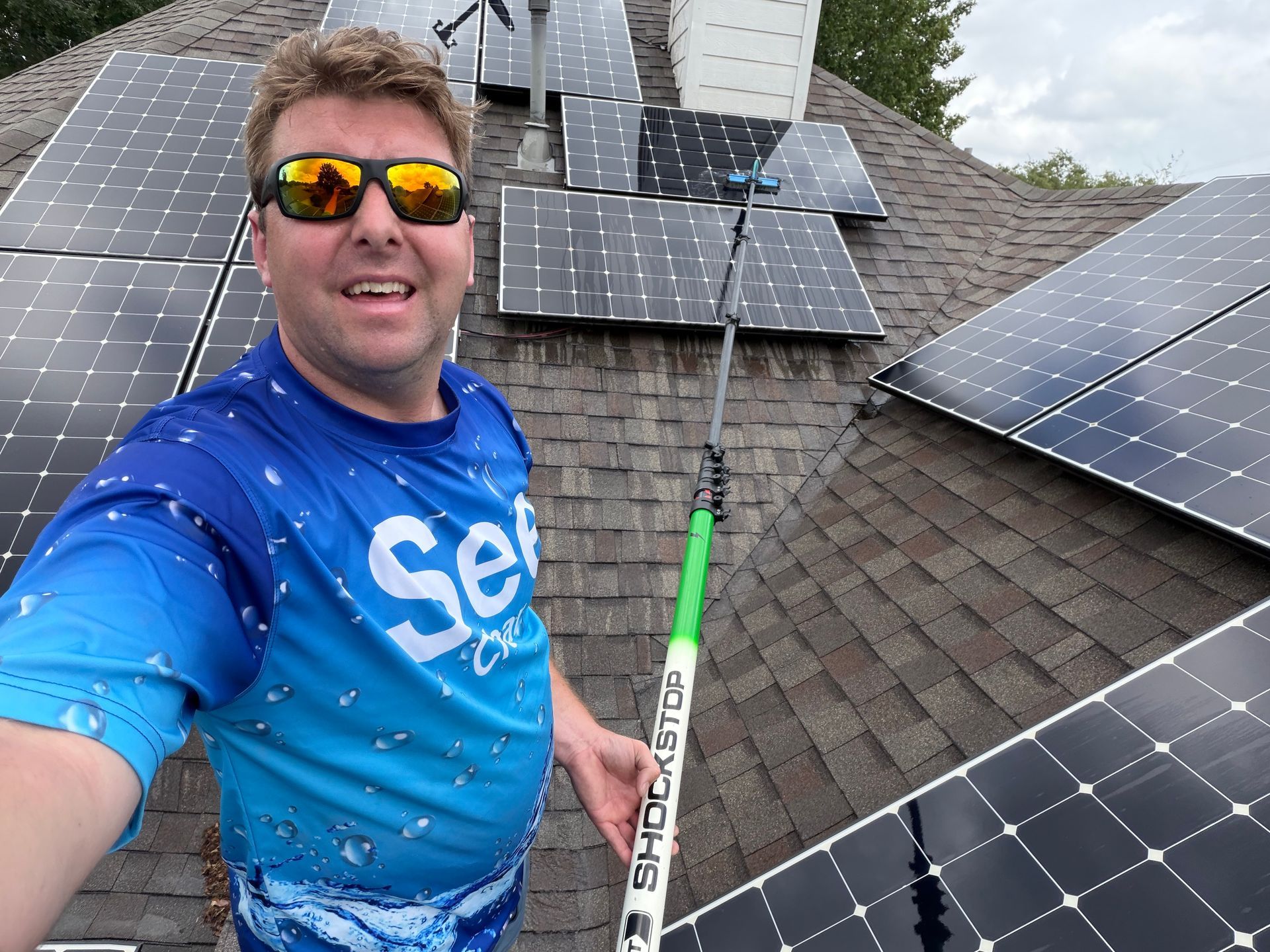 Man wearing sunglasses smiling on a solar-panel roof, holding a cleaning pole.