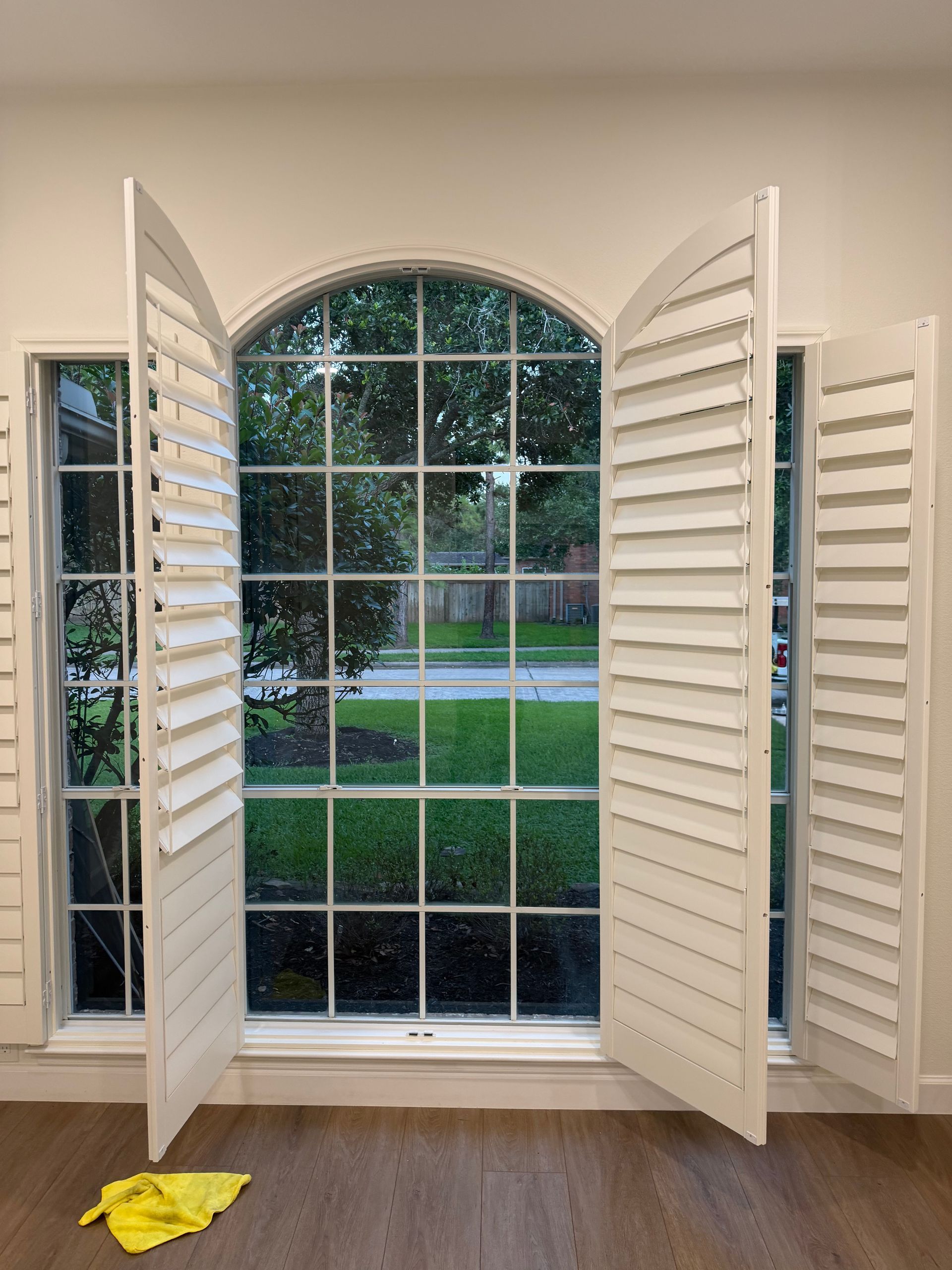 Open white shutters framing an arched window with a view of a green yard and street outside.