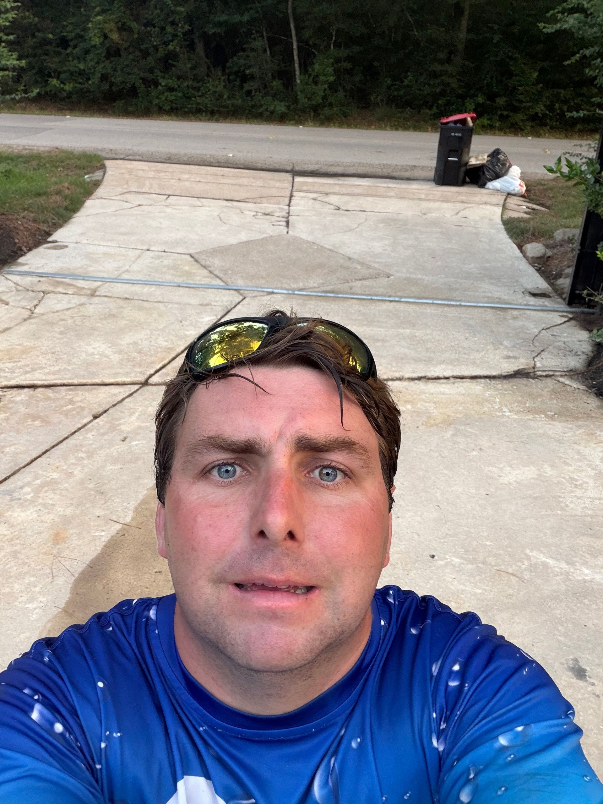 Selfie of a man in a blue shirt on a driveway, with sunglasses on his head and a trash bin in the background