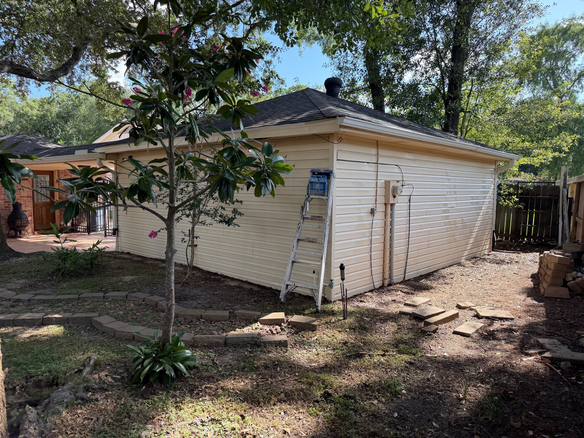 White house exterior with a ladder against the wall, surrounded by trees and a dirt yard.
