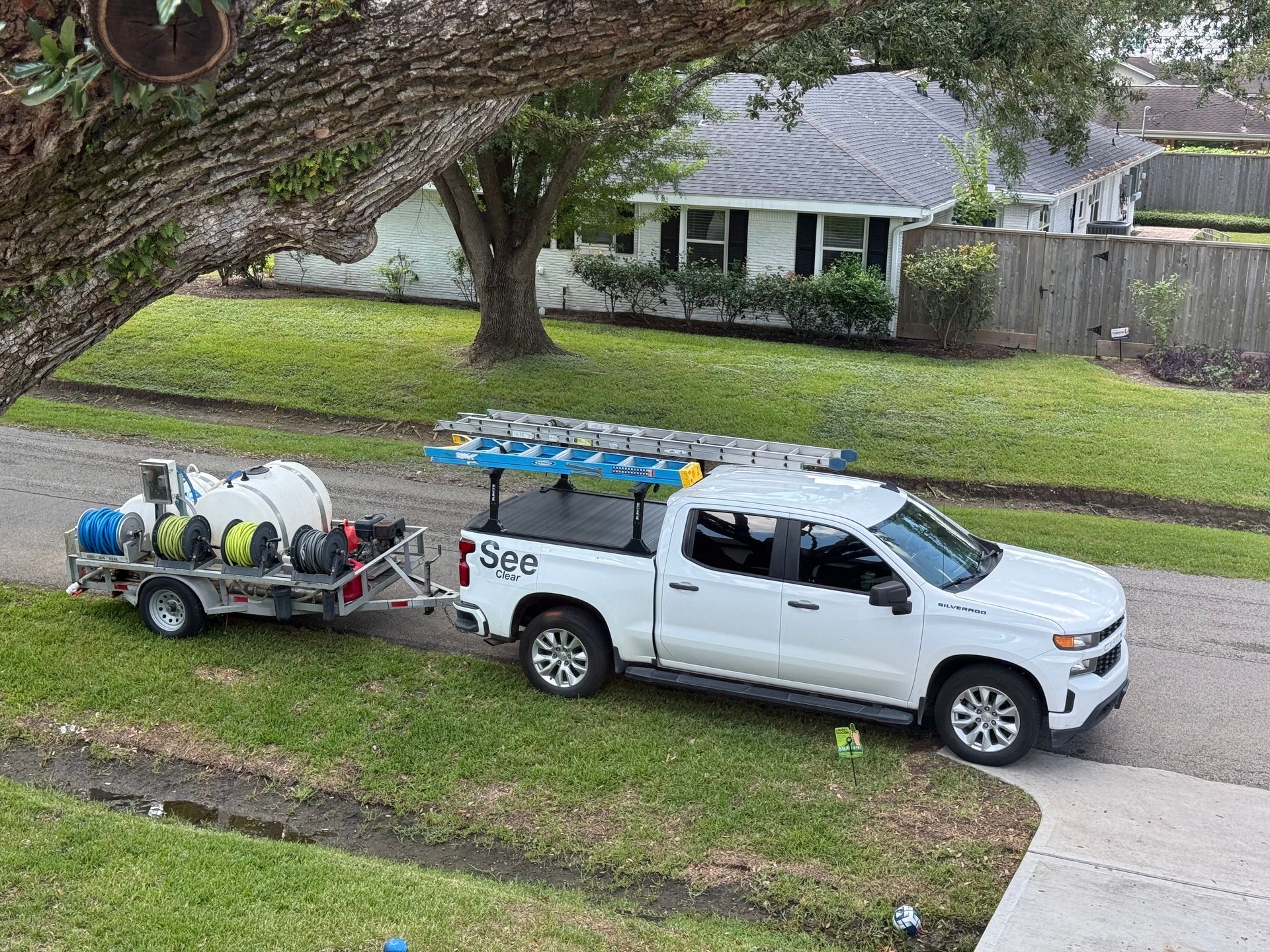 White service truck towing a hose trailer on a suburban street, with large tree and houses in the background