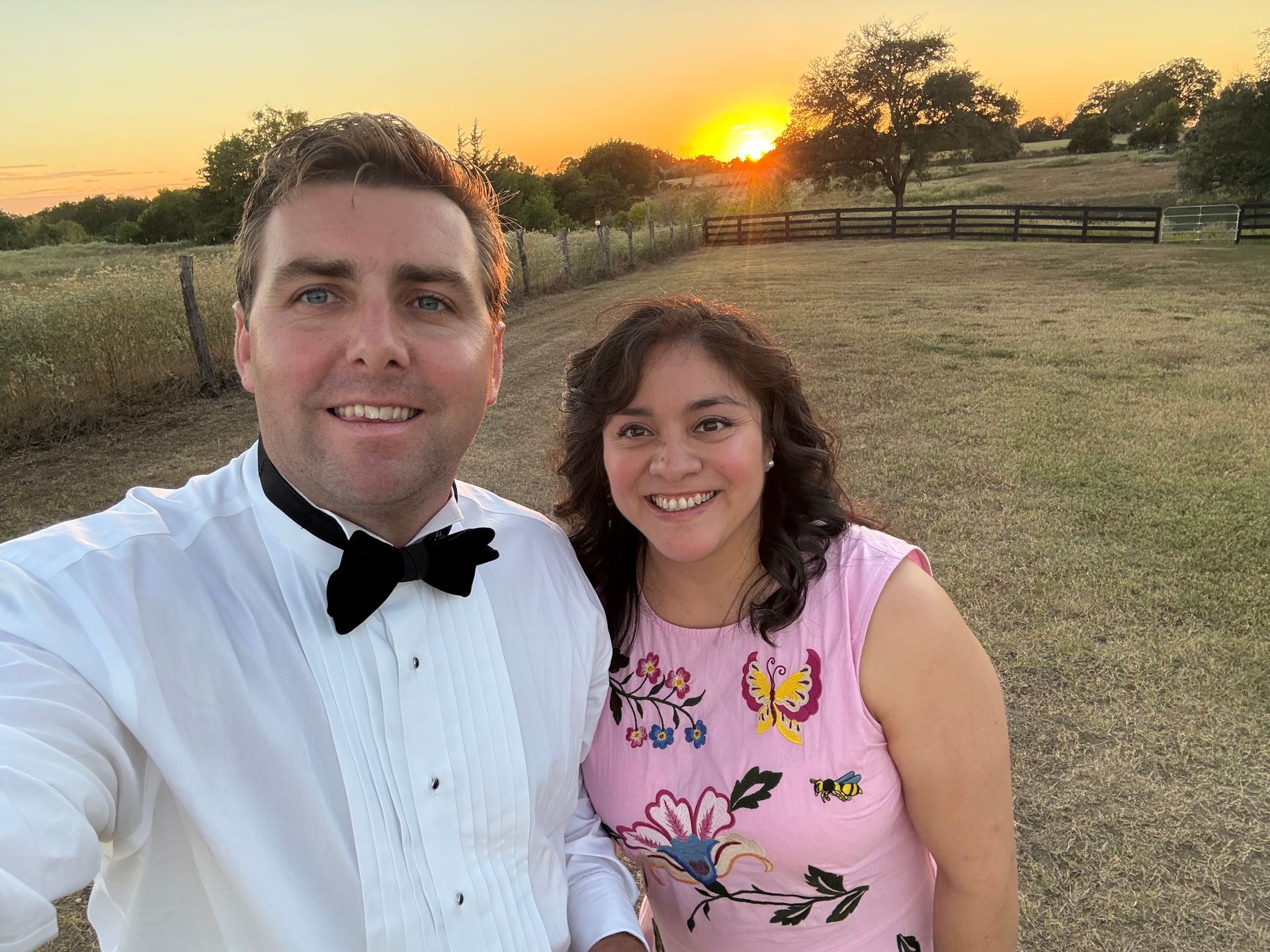 Couple smiling outdoors at sunset, with grassy field and fence in the background.