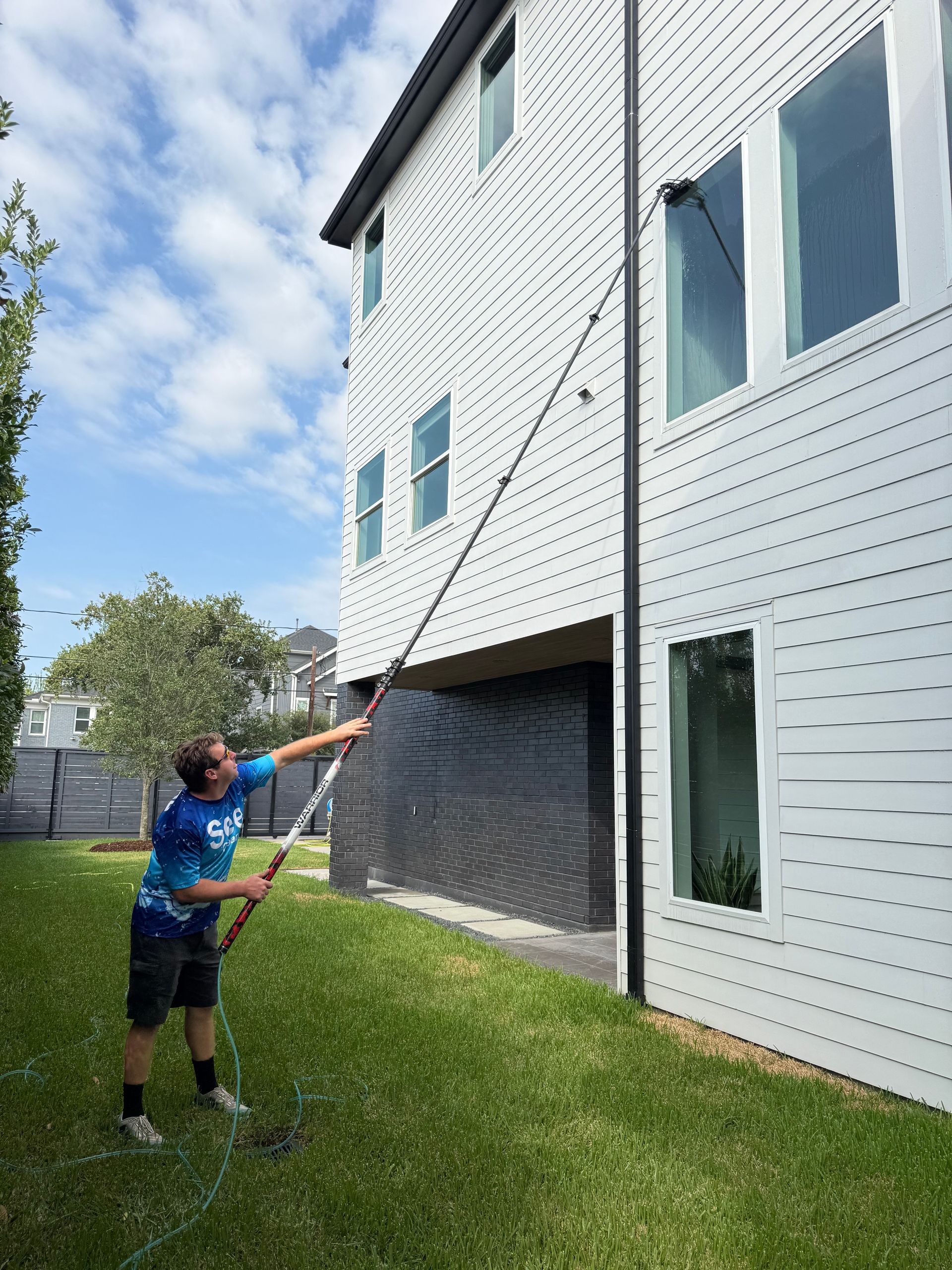 Person using a long-handled tool to clean a white house exterior beside a lawn and driveway