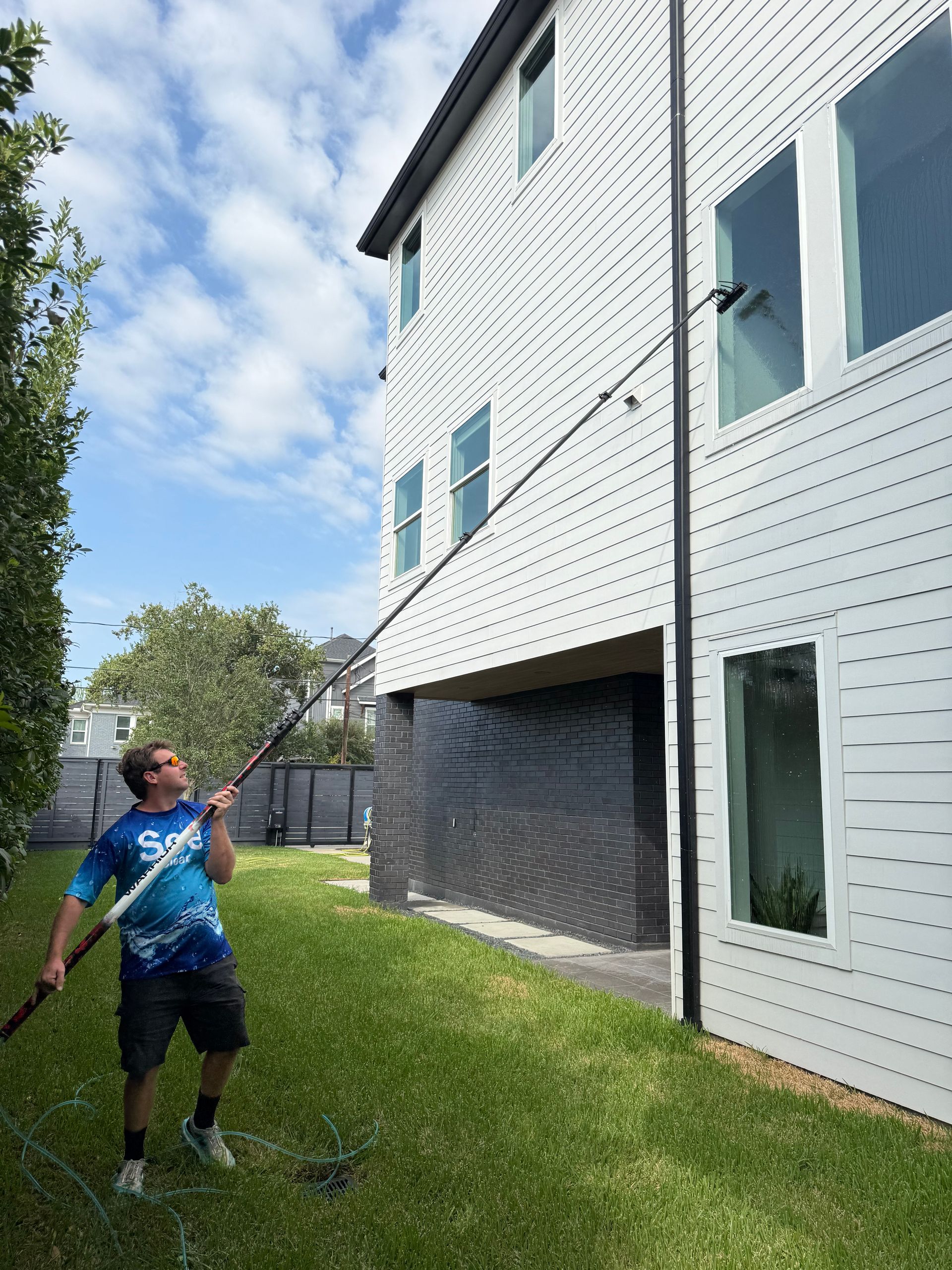 Person standing on grass beside a white house, holding a hose under a blue sky.