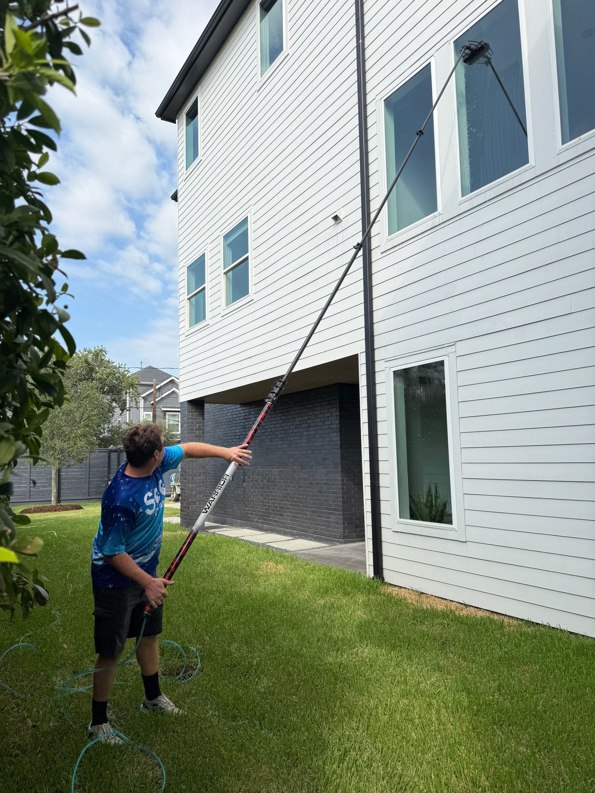 Person using a long pole to reach a second-story window beside a white house and green lawn
