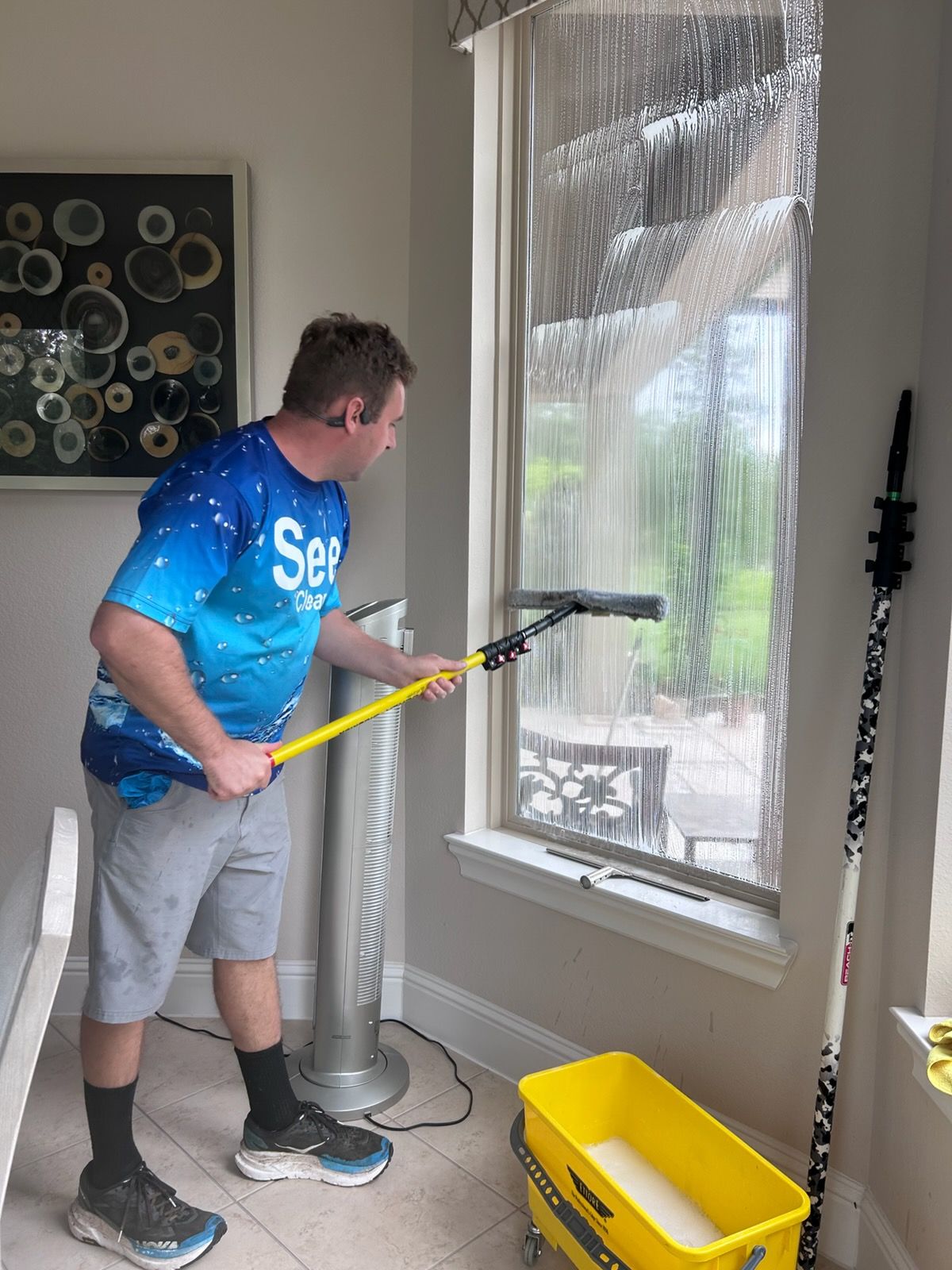 Person squeegeeing a window indoors, with a yellow bucket and plastic sheeting nearby.