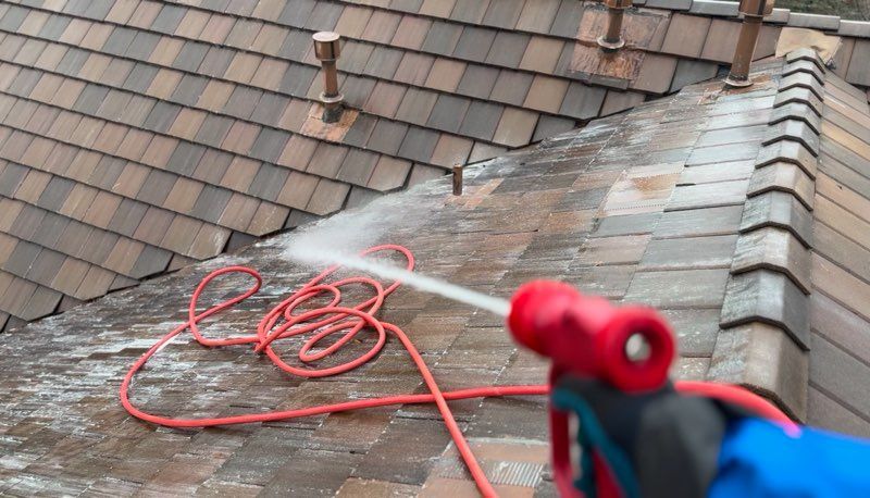 Roof being power-washed, with a red hose and spray nozzle visible on the shingles.