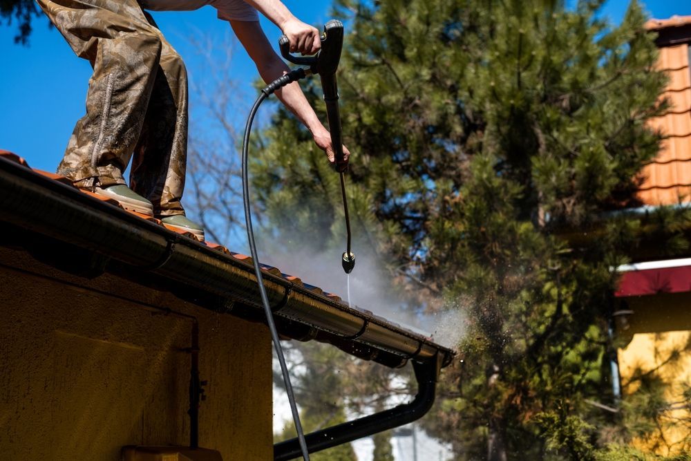 Person using a pressure washer on a roof gutter beside a house, with water spraying into trees