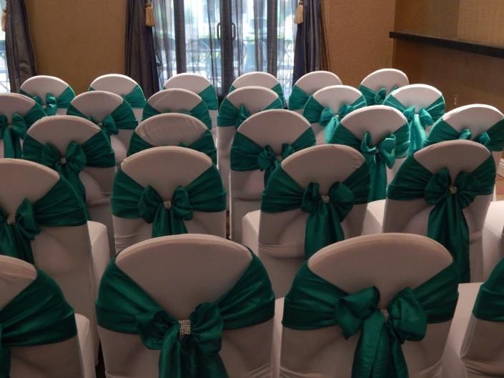 Rows of chairs with white covers and dark green bows set up in a decorated event hall.