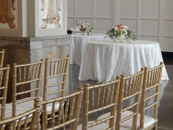 White-draped table with flower arrangements beside rows of gold chairs in a decorated hall