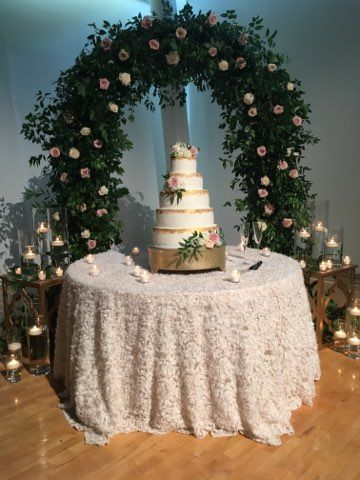 Elegant wedding cake on a lace-covered table beneath a floral arch with candles on either side.