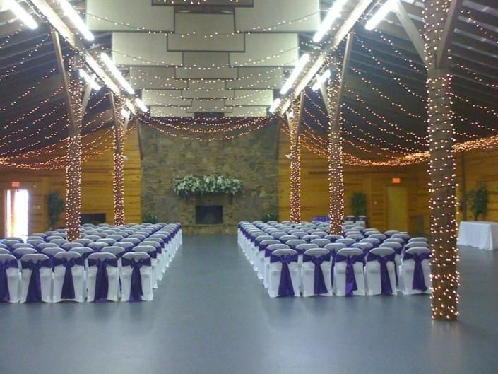 Decorated banquet hall with rows of white-covered chairs and twinkling string lights around pillars