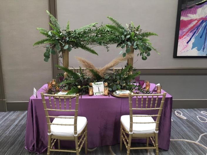 Decorated banquet table with purple tablecloth, tropical greenery centerpiece, and gold chairs in a hotel hallway