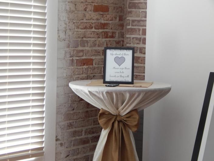 Small round table with white drape and gold bow, brick wall backdrop, and a framed sign on top