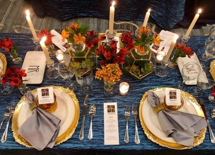 Candlelit dining table set with gold plates, gray napkins, flowers, and place cards on a blue textured tablecloth