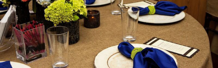 Dining table set with white plates, blue napkins, clear glasses, and yellow flowers.