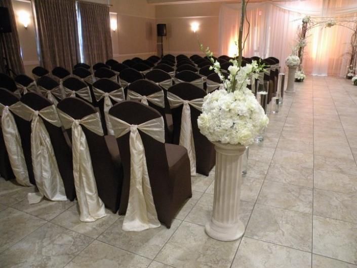 Decorated event hall with rows of black chairs, cream sashes, and white floral arrangements on pedestals
