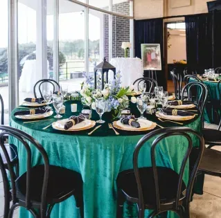 Round banquet table set with black chairs, green tablecloth, floral centerpieces, and lanterns in a bright room