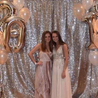 Two women posing in front of a silver sequin backdrop with gold and white balloons