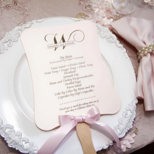 Pink wedding program on a white lace plate with a pink ribbon and wooden handle, on a blush table setting