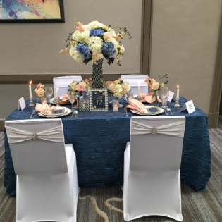 Decorated banquet table with blue and white floral centerpiece, place settings, and two white chairs in a hall