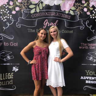Two graduates posing in front of a “Congratulations Grad” backdrop, one in red and one in white.