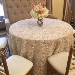 Round lace-covered dining table with a flower centerpiece, surrounded by upholstered chairs in a softly lit room
