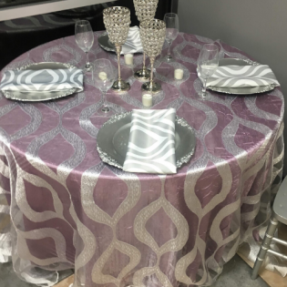 Round dining table set with pink patterned tablecloth, silver placemats, napkins, wine glasses, and centerpiece candles