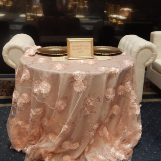 Small round table draped in blush pink lace cloth with a framed sign, set in a dim lounge.